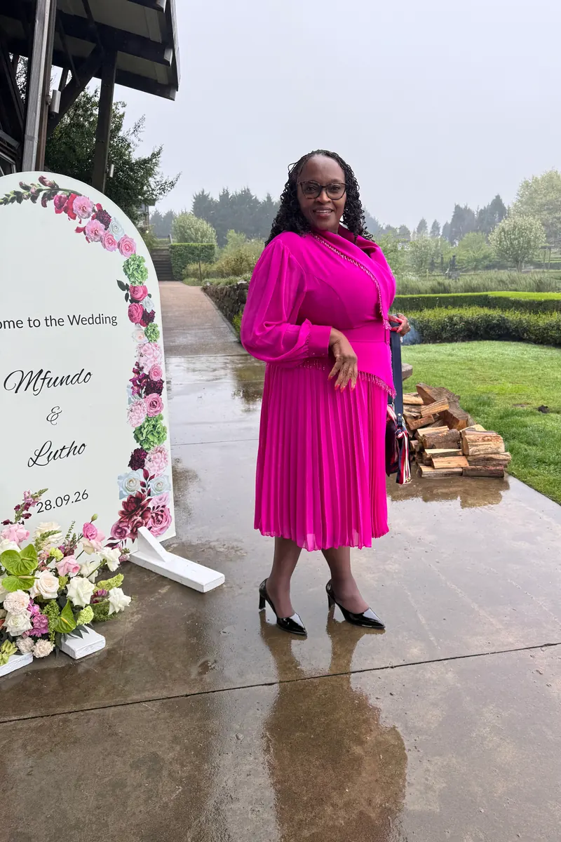 A wedding guest in a pink dress beside a floral welcome sign on a rainy terrace, raw phone photo