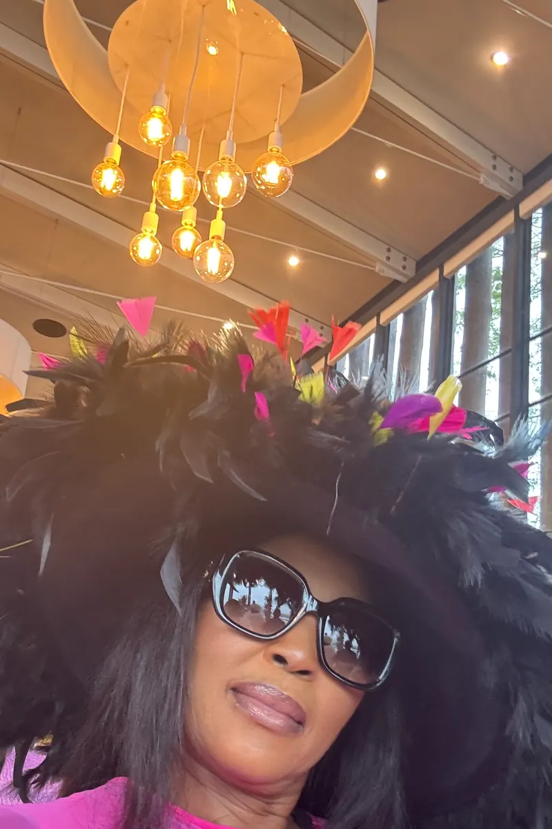 A wedding guest in a feathered hat and sunglasses taking a selfie under chandeliers, raw phone photo