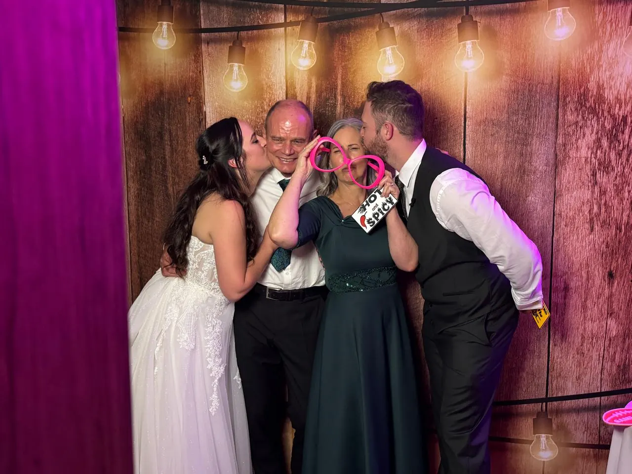A bride and groom kissing their parents in front of a rustic wood and hanging-light backdrop at a KZN Midlands wedding